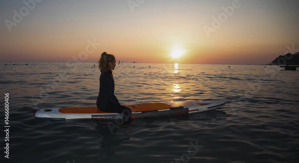 Fototapeta An elderly woman rides paddleboarding in the ocean. Senior female is waiting for a wave on a surfboard. Active old age. Old girl is sitting on a board in the sea at sunset, dressed in a swimming suit