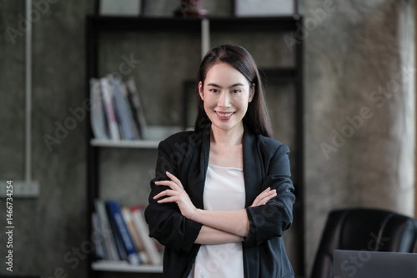 Fototapeta A confident asian businesswoman stands with arms crossed in a modern office, wearing a blazer and smiling professionally at the camera.