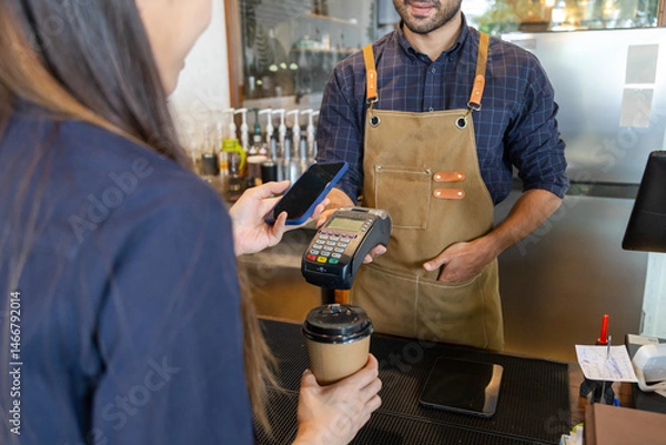 Fototapeta Young Indian baristas or cashier workers man holding a payment machine for scan, cashless technology and credit card payment concept. Customer using smartphone for payment to owner at cafe restaurant