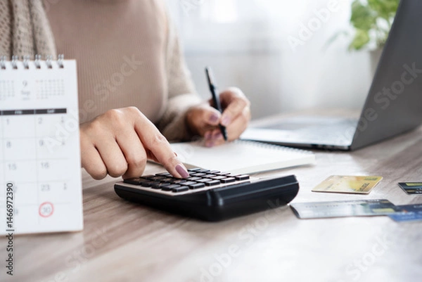 Obraz Woman calculating monthly debt payments, writing down numbers with a calendar with marked due dates, using a calculator with  credit card on the desk