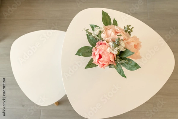 Fototapeta Top-down view of a bouquet of artificial pink peonies and green foliage placed on a pair of white, modern nesting tables. T