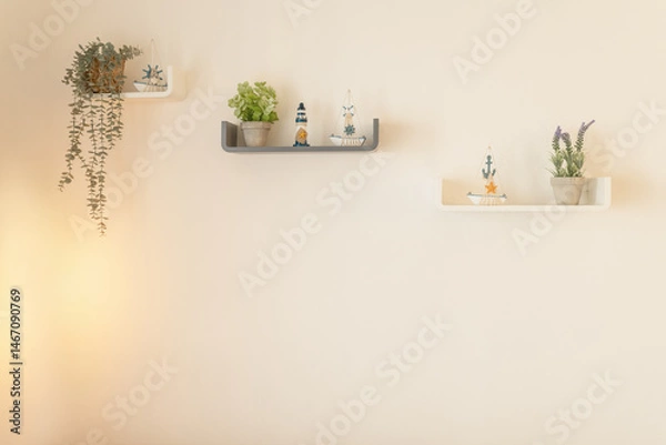 Fototapeta Minimalist wall shelves decorated with small potted plants and nautical-themed ornaments against a soft beige wall. The arrangement includes trailing greenery, a lighthouse figure, and copy space.