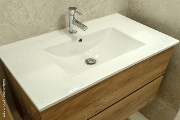 Fototapeta Modern white ceramic bathroom sink with wooden vanity cabinet and chrome faucet. Contemporary and clean design in a tiled bathroom setting.