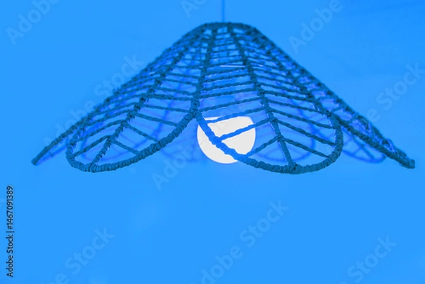 Fototapeta Close-up of a ceiling lamp with a flower-shaped woven lampshade. The blue lighting creates a cozy and inviting atmosphere.