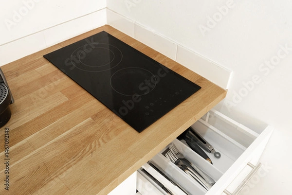 Fototapeta Modern kitchen drawer neatly organized with clean white plates, glass cups, and wine glasses, under a wooden countertop with an induction cooktop. A tidy and stylish kitchen space close up.