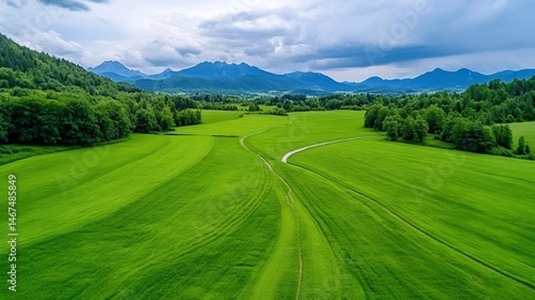 Obraz Aerial View Of Lush Green Field Landscape With Winding Pathway And Mountain Range Background