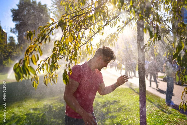 Fototapeta A happy young man stands in a sunny park, smiling with eyes closed as he enjoys the refreshing water spray from active sprinklers
