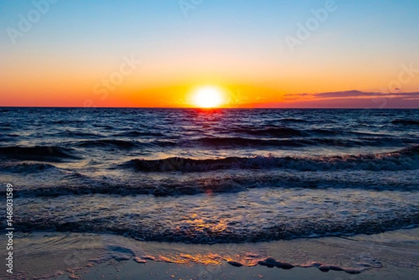 Fototapeta A breathtaking ocean sunset with the sun dipping below the horizon, casting a warm golden glow on the rippling blue water. Serene and picturesque coastal scene.