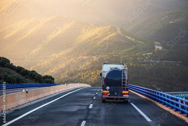 Fototapeta Tanker truck driving on a highway at dawn, with mountains and a forest in the background, rear view.