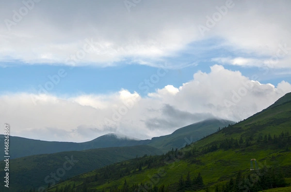 Fototapeta Serene mountain scenery featuring layered hills shrouded in clouds under a bright blue sky. A tranquil atmosphere of natural beauty and calmness, perfect for themes of escape and nature's serenity