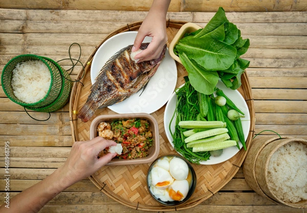 Fototapeta A traditional Thai meal is beautifully arranged on a round bamboo tray. At the center is a perfectly grilled whole fish, served alongside a variety of fresh vegetables, cucumbers, and boiled eggs.