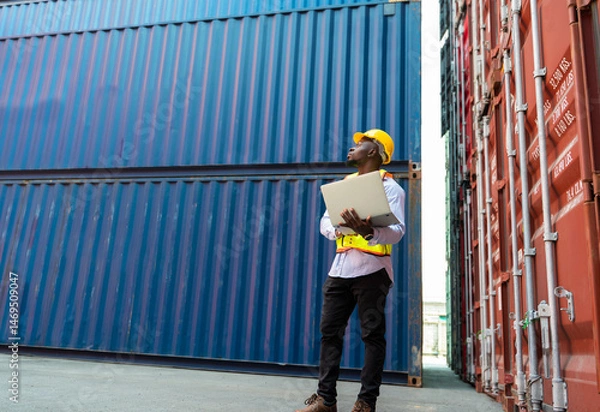 Fototapeta African engineer in yellow hard hat and safety vest holding laptop. Foreman looking to blue shipping container for loading inspection at cargo shipyard.