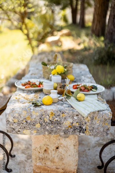 Fototapeta A Mediterranean meal is served on a weathered stone table under the trees with lemons and olive oil. This image captures slow living and rustic elegance perfect for food lifestyle or travel content