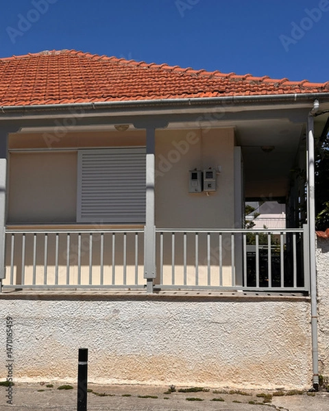 Fototapeta Exterior of a classic house with red tiled roof, closed white shutters, and railing, under clear blue sky, captured in natural daylight