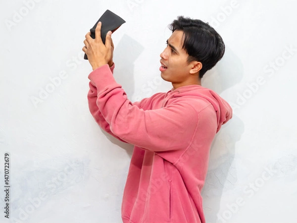 Fototapeta Poor Young Asian man wearing a pink hoodie looking sad and frustrated while holding an empty wallet. Isolated on a white background. Perfect for concepts of poverty or financial struggle