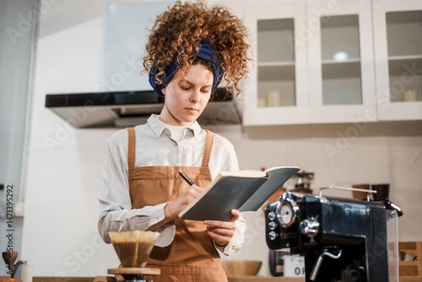 Obraz A young woman with curly hair and a headband, wearing a brown apron, prepares pour-over coffee on a wooden countertop in a bright kitchen. Focused and calm, she enjoys her morning routine.