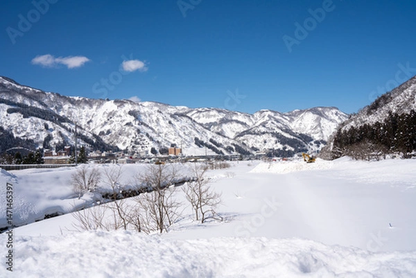 Fototapeta A bright yellow excavator works atop a snow pile in a serene winter valley, surrounded by snow-covered fields, pine forests, and majestic mountains under a vibrant blue sky.
