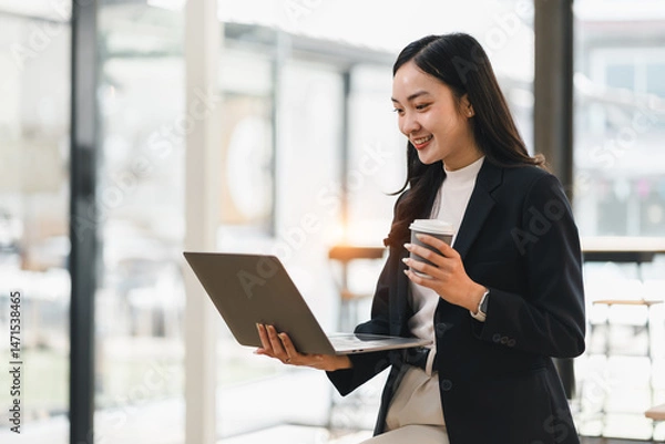 Fototapeta Young Asian businesswoman, smiling and holding coffee cup, works on laptop in modern office with bright natural light, expressing confidence and productivity