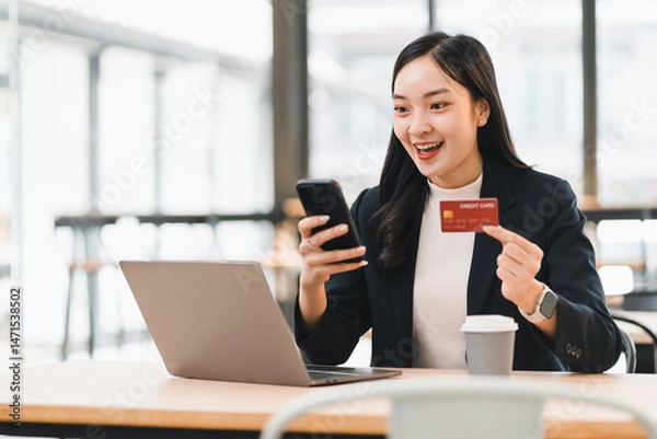 Fototapeta Asian woman business attire holding credit card using smartphone sitting at table laptop coffee cup bright office online shopping happy expression