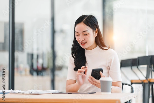 Fototapeta Young Asian woman sitting at desk in modern office, smiling while using smartphone and holding coffee cup, with documents and tablet on table, natural light ambiance