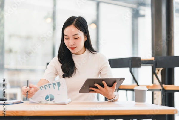 Fototapeta Young Asian businesswoman working with financial chart, holding tablet and writing notes at modern office, focused and professional