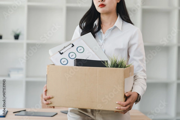 Fototapeta Woman holding cardboard box with office supplies documents and plant standing in modern office concept of job loss resignation or career change feeling uncertain and anxious
