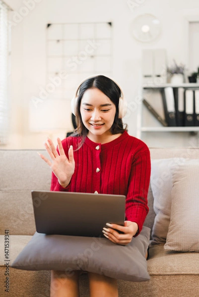 Fototapeta Young Asian woman wearing headphones and red sweater sits on sofa with pillow, smiling and waving during video call on laptop in cozy living room