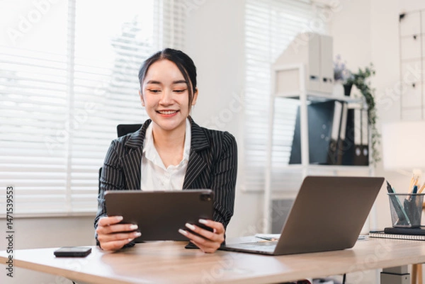 Fototapeta Young Asian businesswoman, smiling and confident, sits at modern office desk using digital tablet, with laptop and smartphone, natural daylight through window blinds