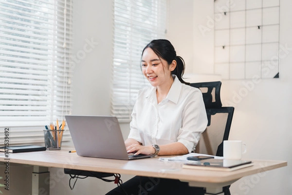Fototapeta Young Asian woman working at modern office desk, smiling while using laptop, surrounded by documents, coffee cup, and stationery, in bright workspace with natural light