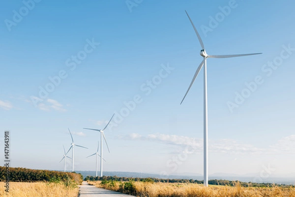 Fototapeta Amidst a winter meadow windmill farm turbines harness the wind's force for sustainable energy. Modern wind technology supporting a cleaner environment under a blue sky.