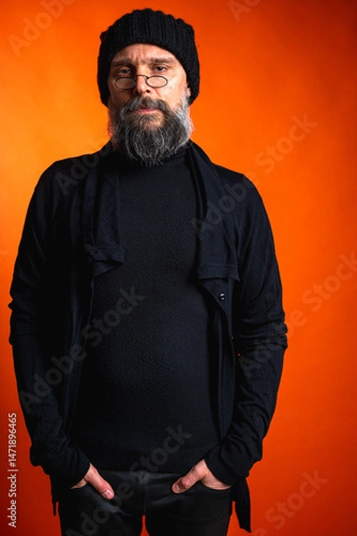 Obraz Portrait of a stylish mature man with a beard and glasses wearing a black hat and clothes, standing against a vibrant orange background. Trendy, confident, modern look.