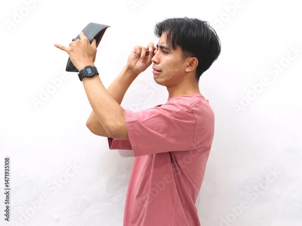 Fototapeta A young Asian man in a pink shirt holds an empty wallet with a sad expression on a white isolated background.