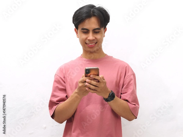 Fototapeta Young Asian man in a pink T-shirt smiling while holding and focusing on a mobile phone against a white background.