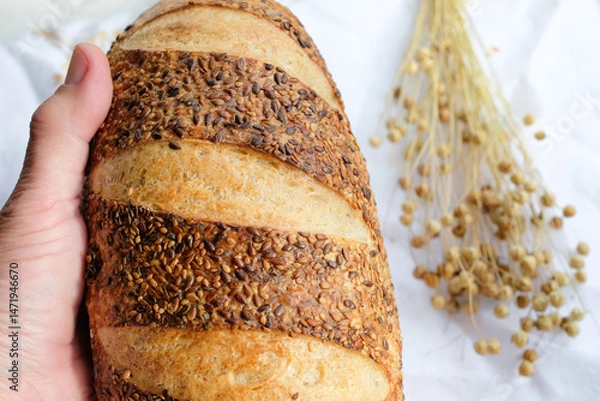 Fototapeta Bread loaf sprinkled with flax and sesame seeds in woman's hand. Flax seed pods. Sourdough bread. Top view. Shallow depth of field.