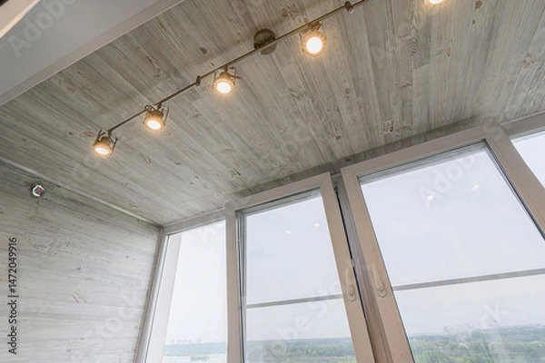 Fototapeta view from indoors showing a light gray, wood-paneled ceiling with track lighting and open windows, overlooking a city skyline