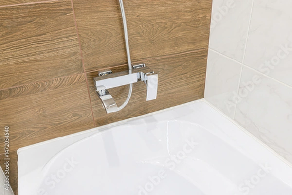 Fototapeta close-up of a bathtub corner with a chrome faucet mounted on a wall tiled with wood-like and white marble