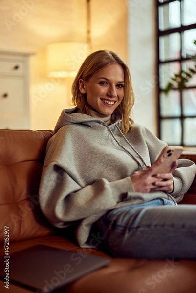 Fototapeta Relaxed Woman Using Smartphone in Cozy Living Room Setting, Smiling at Camera