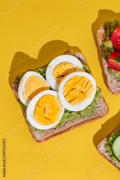 Obraz Delicious healthy bread toasts with fresh avocado, boiled eggs, cherry tomato and cucumber placed on bright yellow background