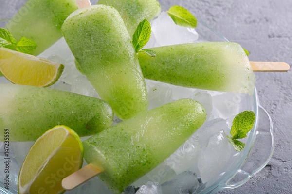 Fototapeta Homemade lime and mint leaves popsicles piled up on a plate of ice on grey concrete background. Side view
