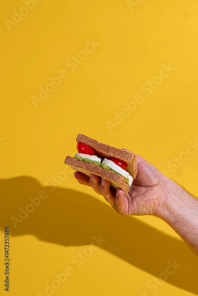 Obraz Man holding delicious sandwich with grilled bread, avocado, boiled eggs, cherry tomato over yellow background. Concept of food, taste, breakfast. Complementary colors. Poster, ad