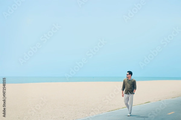 Fototapeta Coastal Stroll: A man strolls along a paved path near a sandy beach, the ocean stretching out before him under a clear sky. The scene evokes feelings of serenity and freedom.