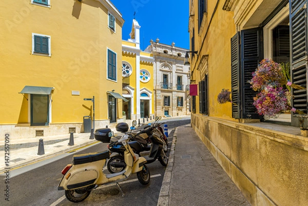 Fototapeta Narrow street with scooters parked against a backdrop of vibrant yellow buildings and historic architecture in Mahon, Spain.