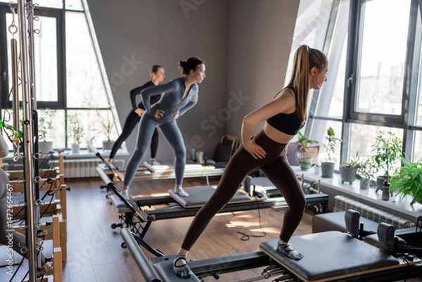 Fototapeta Young caucasian women exercising on pilates reformers in a bright studio. Their workout focuses on core strength and flexibility in a modern fitness environment.