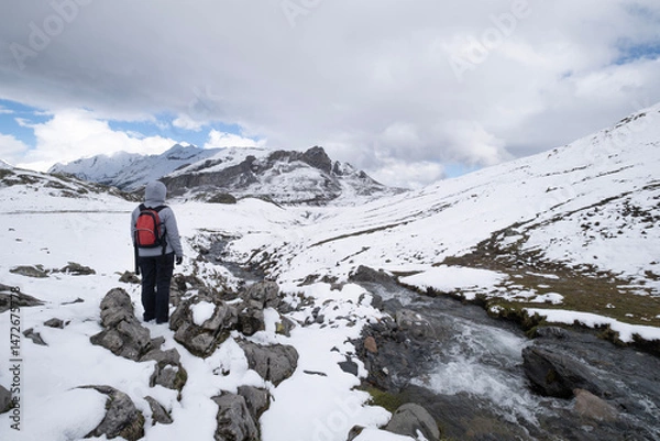 Obraz Hiker with a red backpack stands on snowy rocks, overlooking a mountain stream and snow-covered peaks and a mountain river under a dramatic sky in the Pyrenees