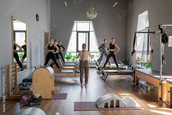Fototapeta Young caucasian women exercising on pilates reformers in a bright studio. Their workout focuses on core strength and flexibility in a modern fitness environment.