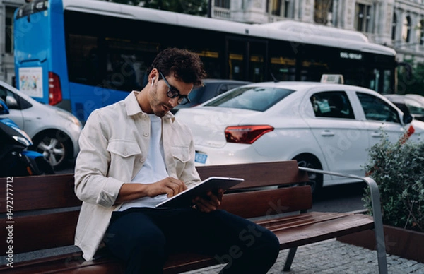 Fototapeta young entrepreneur in stylish attire engages with tablet while sitting on bench near busy street. Focused and determined, he embraces the flexibility of remote work in a fast-paced urban environment