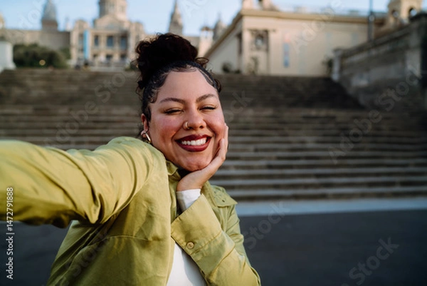 Fototapeta cheerful traveler smiles brightly while taking selfie, capturing joy against backdrop of historic architecture. Female relaxed pose and natural expression reflect a moment of happiness and adventure