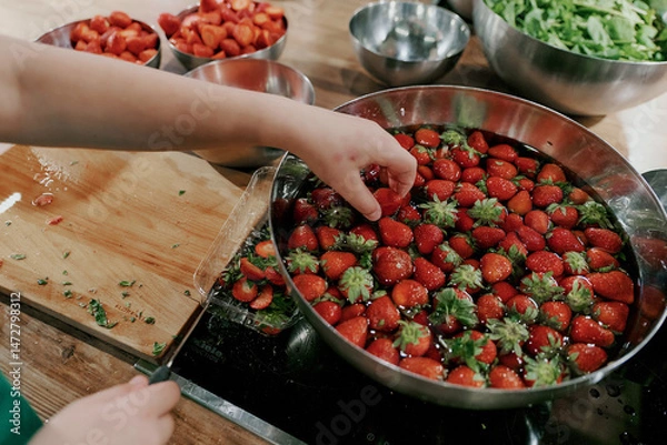 Fototapeta Child’s hand reaching into a bowl of strawberries soaking in water. Surrounded by chopped greens, knife, and small bowls.
