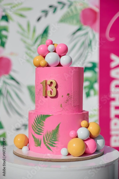 Obraz A vibrant pink birthday cake with a golden number 13, tropical leaf patterns, and colorful edible balls.