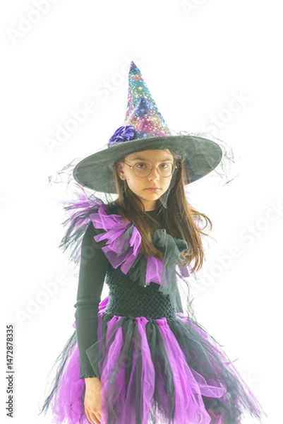 Obraz Cute girl in a witch costume and glasses poses standing in front of a large bright panoramic window. Concept of a children's party and childhood